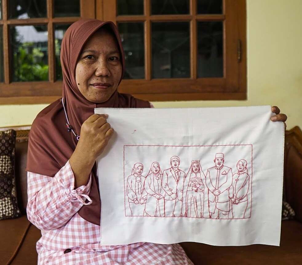 A woman, Ibu Dwi Restu, holds up an embroidery of a family wedding, done in read thread on plain white fabric.