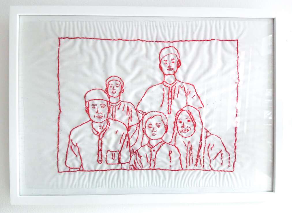 An framed embroidery of Ibu Marini's family. The embroidery is done in red thread on plain white fabric.